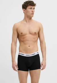 Golu muškarac s kratkom kovrčavom kosom u crnim boksericama s bijelim pojasom marke "Jack & Jones", gleda udesno.