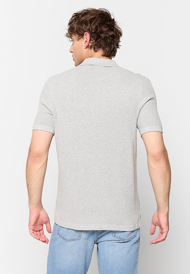 Man met krullend bruin haar in een lichtgrijs gestructureerd polo shirt met korte mouwen en lichtblauwe jeans, met de rug naar ons toegekeerd tegen een witte achtergrond.