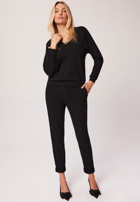 Czarny, prążkowany zestaw loungewear z topem w kształcie V i dopasowanymi spodniami. Posiada boczne kieszenie i podwinięte mankiety, zestawiony z czarnymi szpilkami w kształcie kokardy.