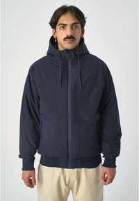 Marineblaue Kapuzenjacke mit einem frontalem Reißverschluss, elastischen Bündchen und seitlichen Taschen, aus leichtem Material gefertigt. Glatte Textur mit minimalistischem Design.