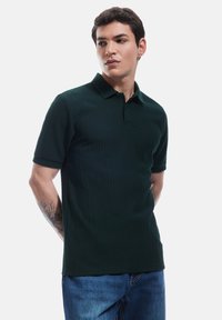Ciemnozielona prążkowana koszulka polo z kołnierzykiem, krótkimi rękawami i prostym dołem, w połączeniu z niebieskimi dżinsami.