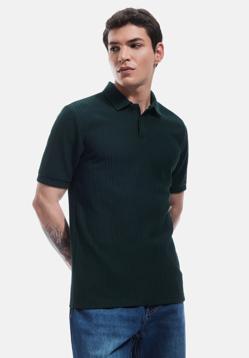 Ciemnozielona prążkowana koszulka polo z kołnierzykiem, krótkimi rękawami i prostym dołem, w połączeniu z niebieskimi dżinsami.