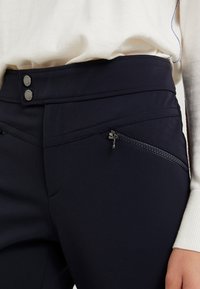 Pantalons de sport bleu marine avec une texture lisse, dotés de deux poches zippées et de coutures contrastantes. Boutons à la taille.