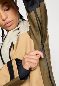 Wasserabweisende Jacke mit Beigen, Braunen und Schwarzen Einsätzen. Sie verfügt über Reißverschlusstaschen und ein strukturiertes Gewebe für Strapazierfähigkeit.