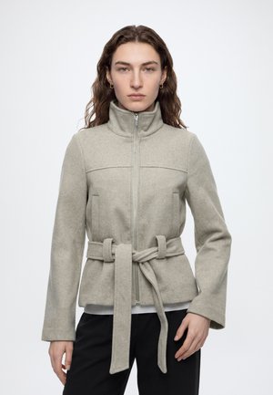 Femme aux longs cheveux bruns portant une veste zippée gris clair avec une ceinture nouée à la taille et un pantalon noir sur un fond uni.
