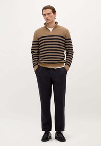 Pull en tricot rayé beige et bleu marine avec un col zippé, associé à un pantalon foncé et des chaussures noires, présentant une coupe décontractée et une texture côtelée.
