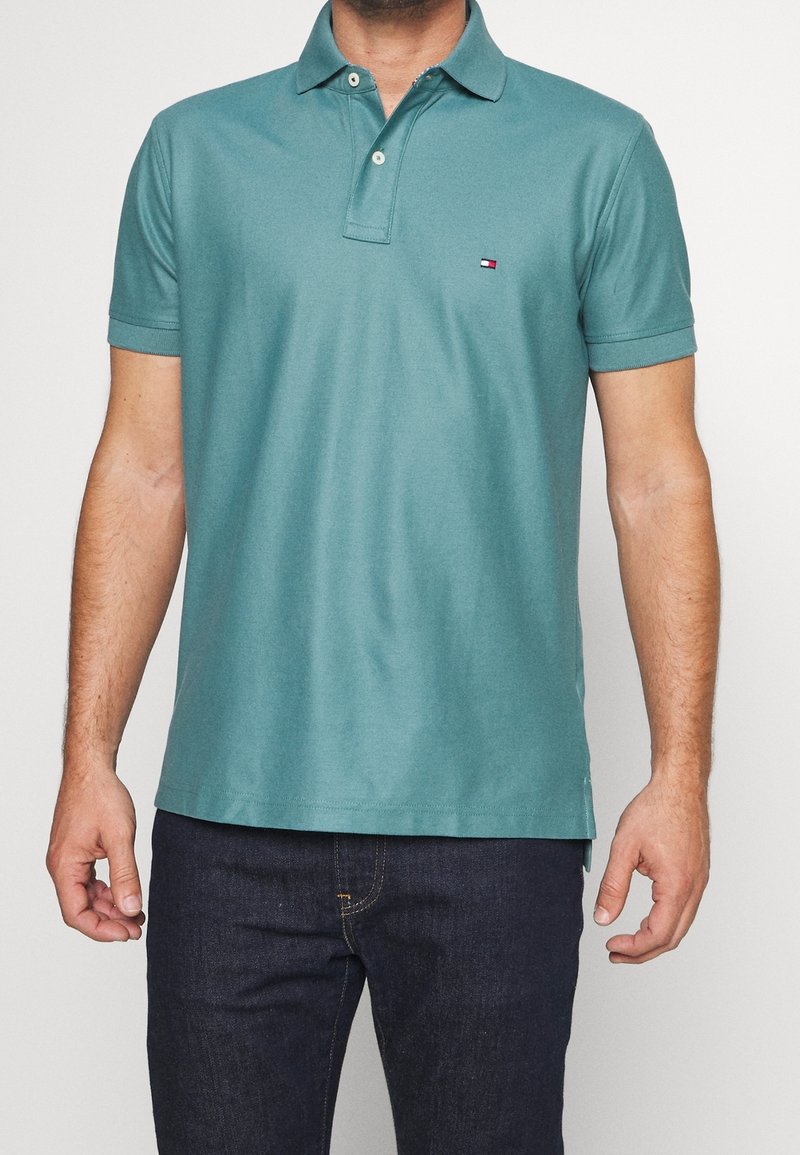 Homme portant un polo bleu sarcelle à manches courtes et un jean foncé, debout avec les bras détendus devant un fond clair uni.