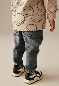 Beige-Hoodie mit schwarzen Smiley-Gesichtern, kombiniert mit grauen konisch zulaufenden Hosen und schwarzen Schuhen mit cremefarbenen Akzenten und elastischen Bündchen.