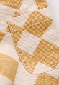 Tissu beige avec un motif texturé subtil présentant de grandes formes géométriques jaunes. Une poche est visible avec une couture contrastante le long du bord.