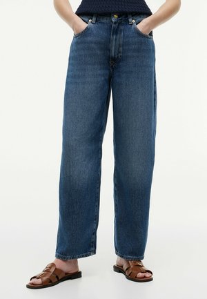 Person trägt locker sitzende blaue Jeans mit den Händen in den Taschen und braune Sandalen mit offenen Zehen vor weißem Hintergrund.