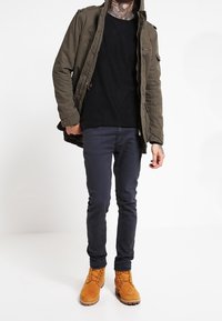 Olivgrüne Parka mit Reißverschluss und Taschen, schwarzes Hemd darunter, navyfarbene schmal geschnittene Hosen und tanfarbene Wildlederstiefel mit Schnürsenkeln und Gummisohlen.