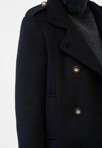 Navy double-breasted coat with gold buttons, notched collar, and a herringbone texture. Accompanied by a dark knit turtleneck.