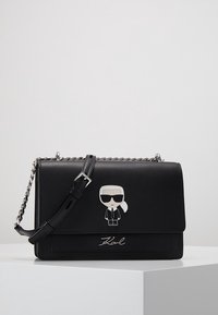 Czarna skórzana torebka z srebrnym łańcuchem, z rysunkową postacią przymocowaną z przodu oraz wytłoczonym logo "Karl" na dole.