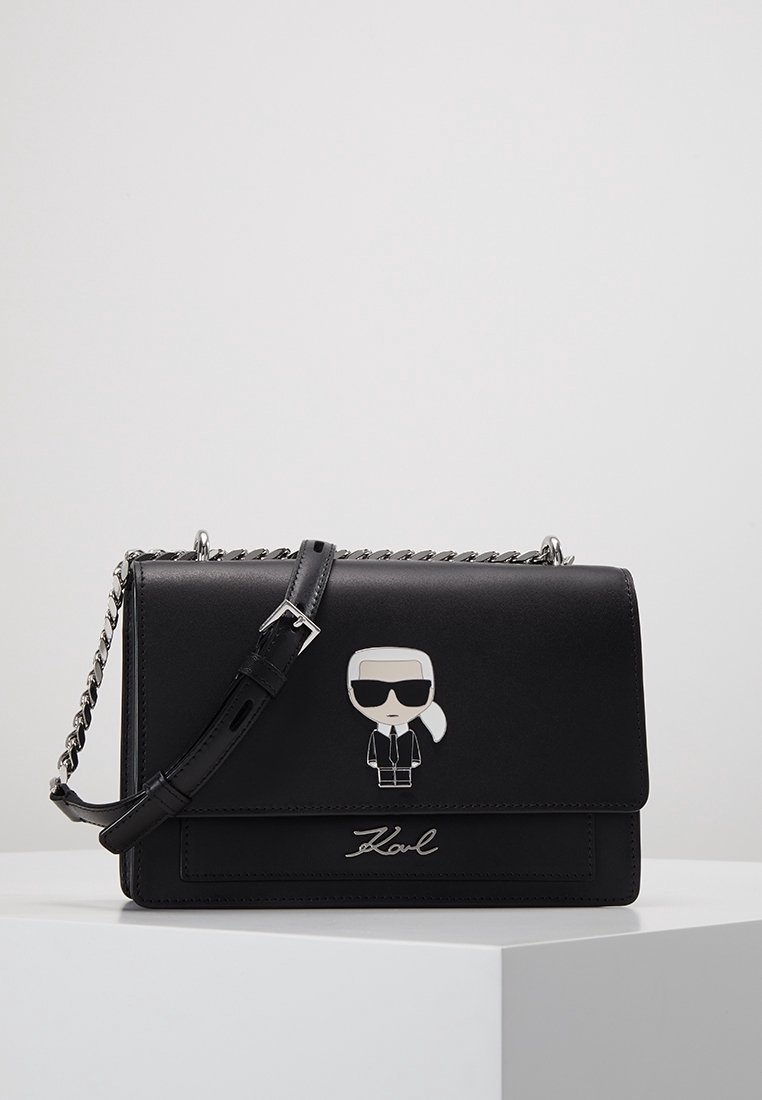 Czarna skórzana torebka z srebrnym łańcuchem, z rysunkową postacią przymocowaną z przodu oraz wytłoczonym logo "Karl" na dole.