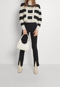 Femme portant un pull court rayé noir et blanc, des leggings noirs avec des fentes à la cheville, des bottes beige, tenant un sac à main blanc à ses côtés.