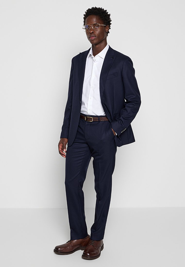 Marineblauer Anzug mit einer taillierten Jacke und passender Hose, kombiniert mit einem weißen Hemd und braunen Lederschuhen, dazu einen braunen Gürtel.