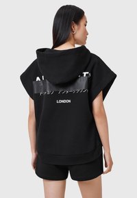 Svart oversized hoodie med korta ärmar, med en texturerad rem över ryggen med vit text och huva. Kombineras med svarta shorts.