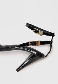 Svart lakklær høyhælte sandaler med to gullspenner og tynne ankel- og tåremmer på hvit bakgrunn.