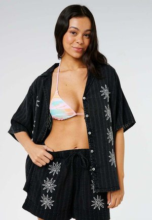 Joven mujer con camiseta negra de manga corta bordada con sol y pantalones cortos sobre un top de bikini de colores pastel, de pie con una mano en la cadera.