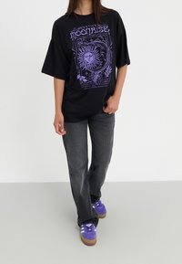 Černé oversized tričko s fialovým grafickým designem, na kterém je slunce a květinové prvky. V kombinaci s tmavě šedými džínami a fialovými teniskami.