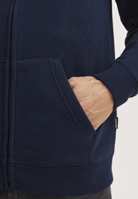 Tumsas zila kapucis jaka ar rāvējslēdzēju, izgatavota no mīksta kokvilnas maisījuma, ar priekšējo kabatu un ribota apakšmalu. Malā ir neliela zīmola birka.