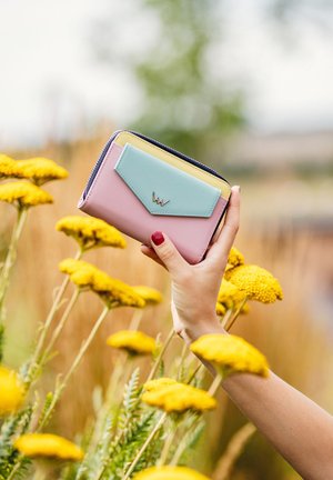 Colorful wallet in pink, mint, and yellow leather, featuring a front flap and a metallic logo, held against vibrant yellow flowers.
