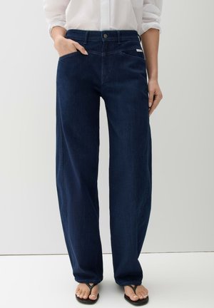 Vrouw draagt donkerblauwe wijde jeans, wit overhemd met lange mouwen opgerold en zwarte teenslippers, staat met één hand in de zak.