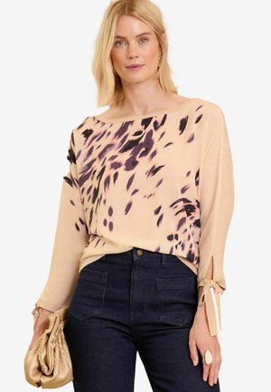Beige top with purple and black abstract patterns, boat neck, long sleeves with tie details, paired with dark jeans and a metallic clutch.