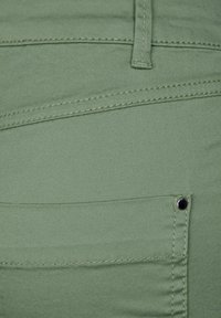 Grön bomullstyg med en slät yta, som har en bakficka med en svart metallknapp och kontrasterande sömdetalj.