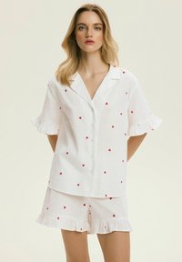 Biały bawełniany zestaw piżamowy z nadrukiem czerwonych serc. Zestaw posiada paski, krótkie rękawy z falbankami oraz koszulę na guziki.