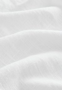 Tissu blanc avec une surface douce et texturée, présentant de fines lignes diagonales et une apparence légère et aérienne. Plissé et superposé.