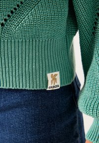 Sweter dzianinowy w kolorze teal z prążkowanym dnem i rękawami. Posiada metkę z logo w złotym kolorze. Założony na ciemnym denimu.