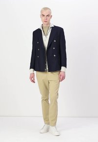 Blazer doppiopetto navy con bottoni metallici, indossato sopra una camicia a fiori chiari, abbinato a pantaloni beige chiari e sneakers bianche.