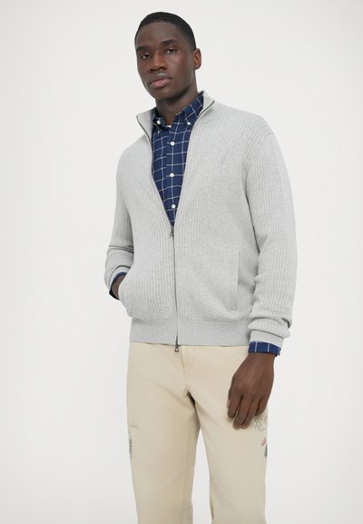 Szürke bordázott, cipzáras kardigán oldalzsebekkel, kék kockás ing fölött viselve. Magas gallérral és mandzsettás ujjakkal rendelkezik.