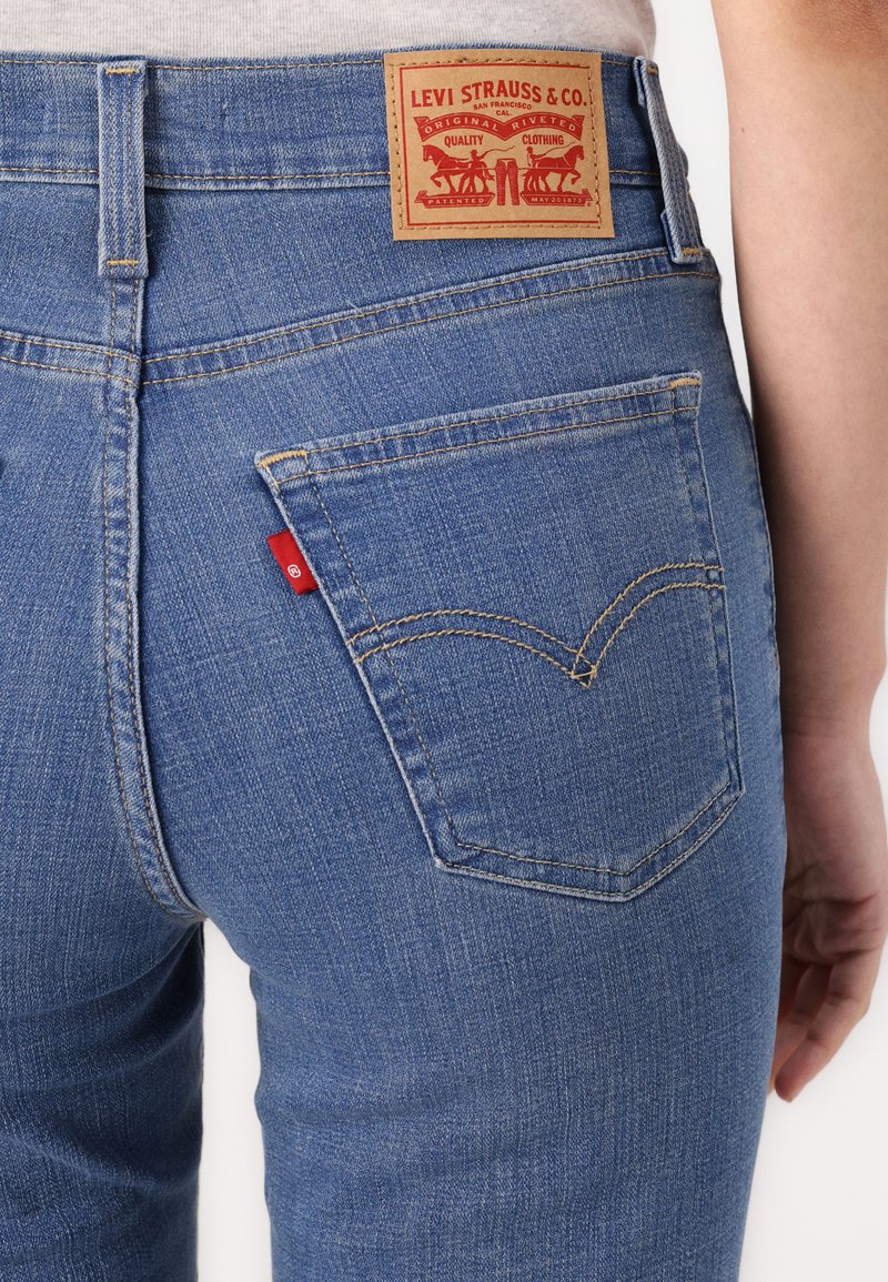 Blaue Denim-Jeans mit hoher Taille, versehen mit einem hellbraunen Lederpatch mit rotem Branding und zwei Gesäßtaschen mit charakteristischen Stickdetails.