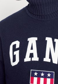 Marineblauer gestrickter Rollkragenpullover mit großen weißen Buchstaben "GAM" und einem rechteckigen patriotischen Aufnäher mit Sternen- und Streifendesign.