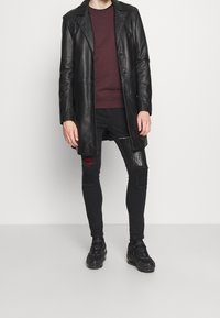 Czarny skórzany płaszcz, bordowa bluza, podarte czarne dżinsy z teksturowanymi akcentami oraz masywne czarne sneakersy. Gładkie tło.