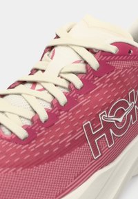 Nærbilde av en rosa og hvit joggesko med hvite lisser og teksturert stoff, med ordet "HOKA" i uthevede bokstaver.