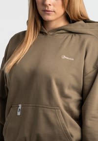 Olivgrön hoodie i mjuk bomull, med en framficka, ribbad mudd och en liten vit logotyp på bröstet. Slät textur.
