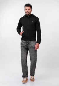 Schwarzer Kapuzenpullover mit Reißverschluss und strukturiertem Muster, mit Kapuze und goldfarbenem Logo. Kombiniert mit grauen Jeans und beigen Turnschuhen.