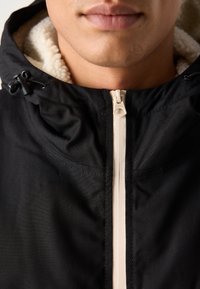 Giacca nera con cappuccio foderato in Sherpa; presenta una zip color crema, regolatori e dettagli di cucitura visibili.