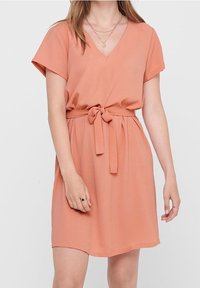 Kortärmad klänning med V-ringning i mjuk persikofärgad tyg, med knytning i midjan, slätt material och avslappnad passform, som slutar ovanför knäet.