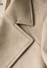 Beżowy wełniany płaszcz z klapami, o gładkiej fakturze i widocznych st seams na brzegach.