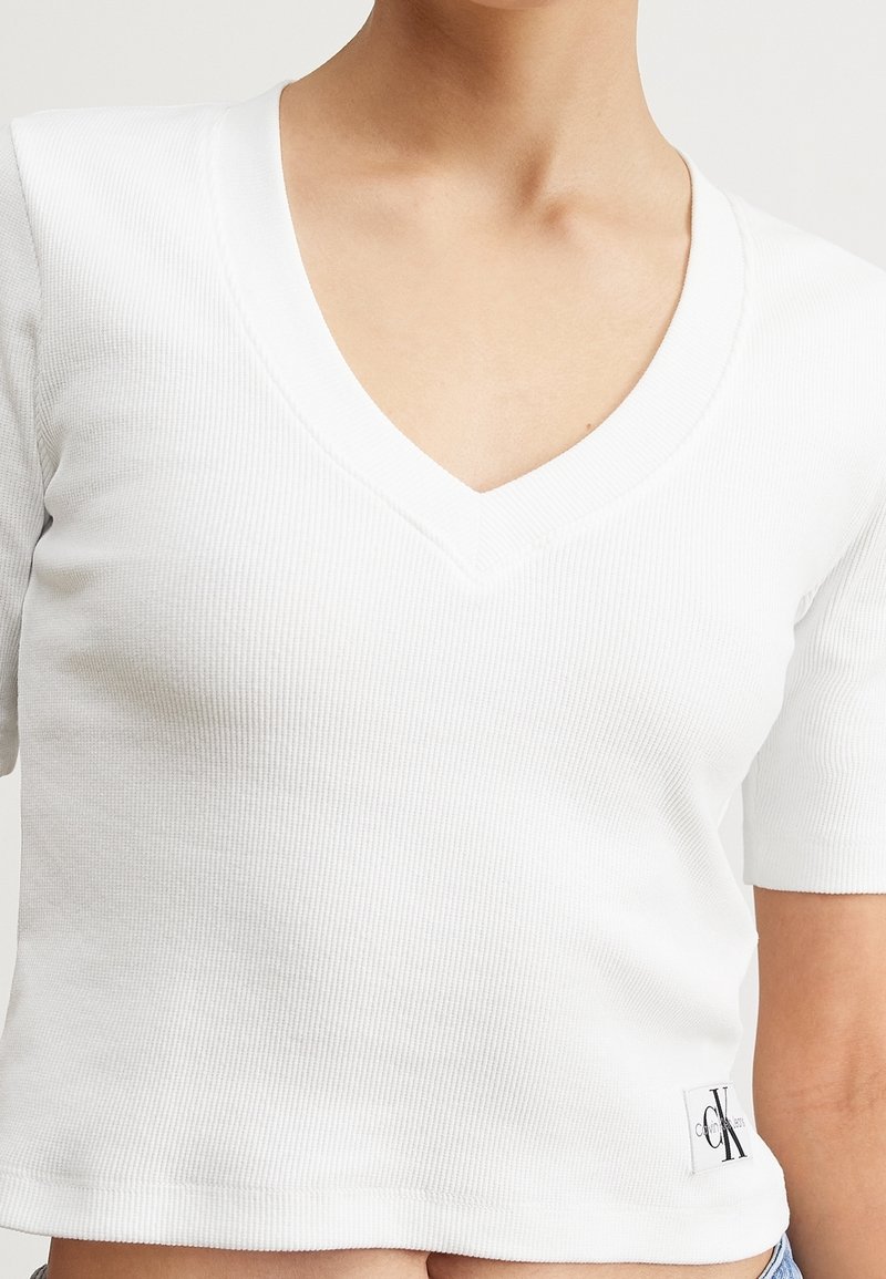 Valkoinen ribattu puuvillainen T-paita V-päällä ja lyhyillä hihoilla. Siinä on pieni brändilogo-patch alaosassa ja sileä pinta.