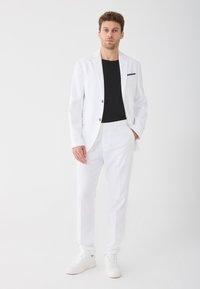 Biały garnitur skrojony na miarę z dopasowanymi spodniami i pasującą marynarką, noszony na czarnej koszulce. Stylizowany z białymi sneakersami. Minimalistyczny design.