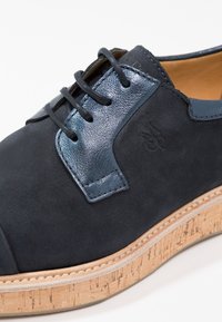 Chaussure en cuir noir avec des accents métalliques bleus, des lacets noirs, un logo embossé sur le côté et une semelle épaisse en liège avec des coutures visibles.