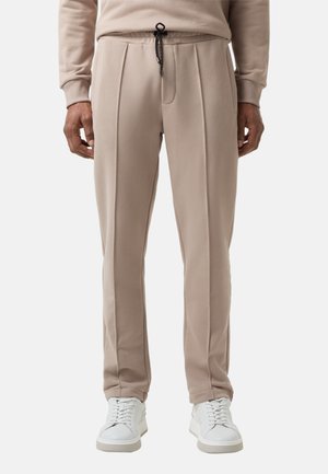 Beige Sweatpants mit lockerer Passform, einem Kordelzugbund, plissiertem Vorderteil und glatter Textur, kombiniert mit weißen Schuhen vor einem schlichten Hintergrund.