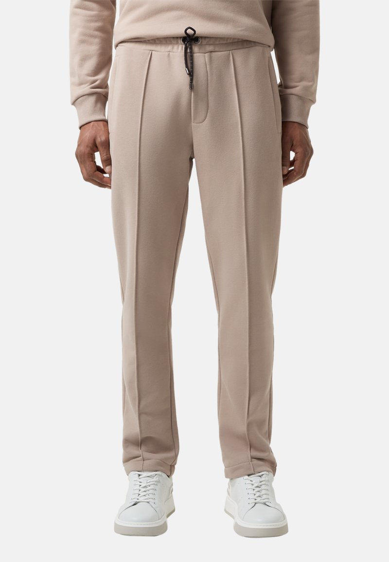 Beige relaxfit joggingbroek met een trekkoord in de taille, geplooide voorkant en een gladde textuur, gecombineerd met witte schoenen op een effen achtergrond.