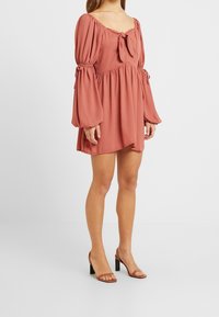 Šaty v rezavé barvě s nabíranými rukávy, detailem na šňůrku u výstřihu a volnějším střihem, doplněné černými sandály na podpatku.