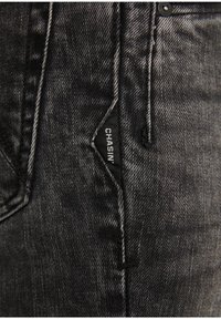 Jeans de denim negro con una textura desgastada, que presentan una etiqueta de logo distintiva, un diseño de cinco bolsillos y cierre de botón metálico.
